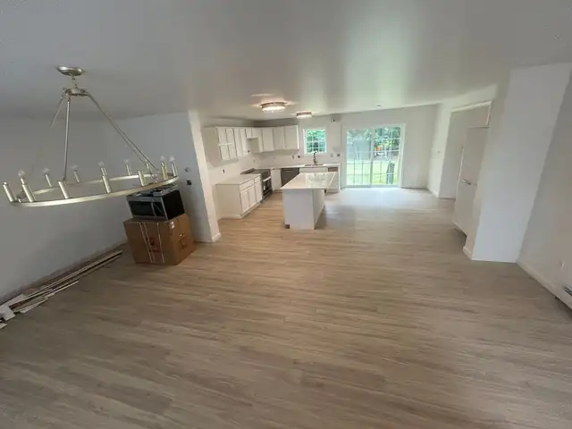 Open kitchen and dining area with white cabinets, island, new plank flooring and a brass chandelier, with a boxed appliance waiting to go in